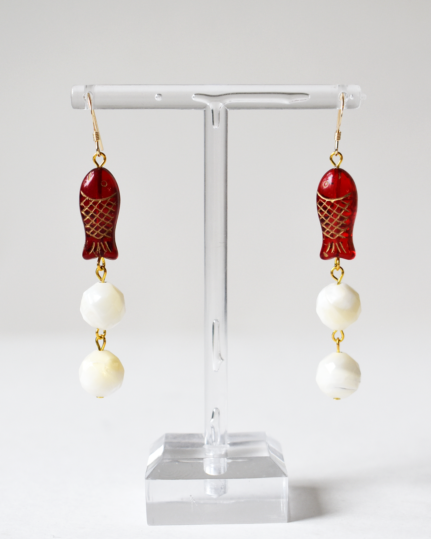 Pair of earrings with red fish-shaped beads and white spherical beads on a clear stand.