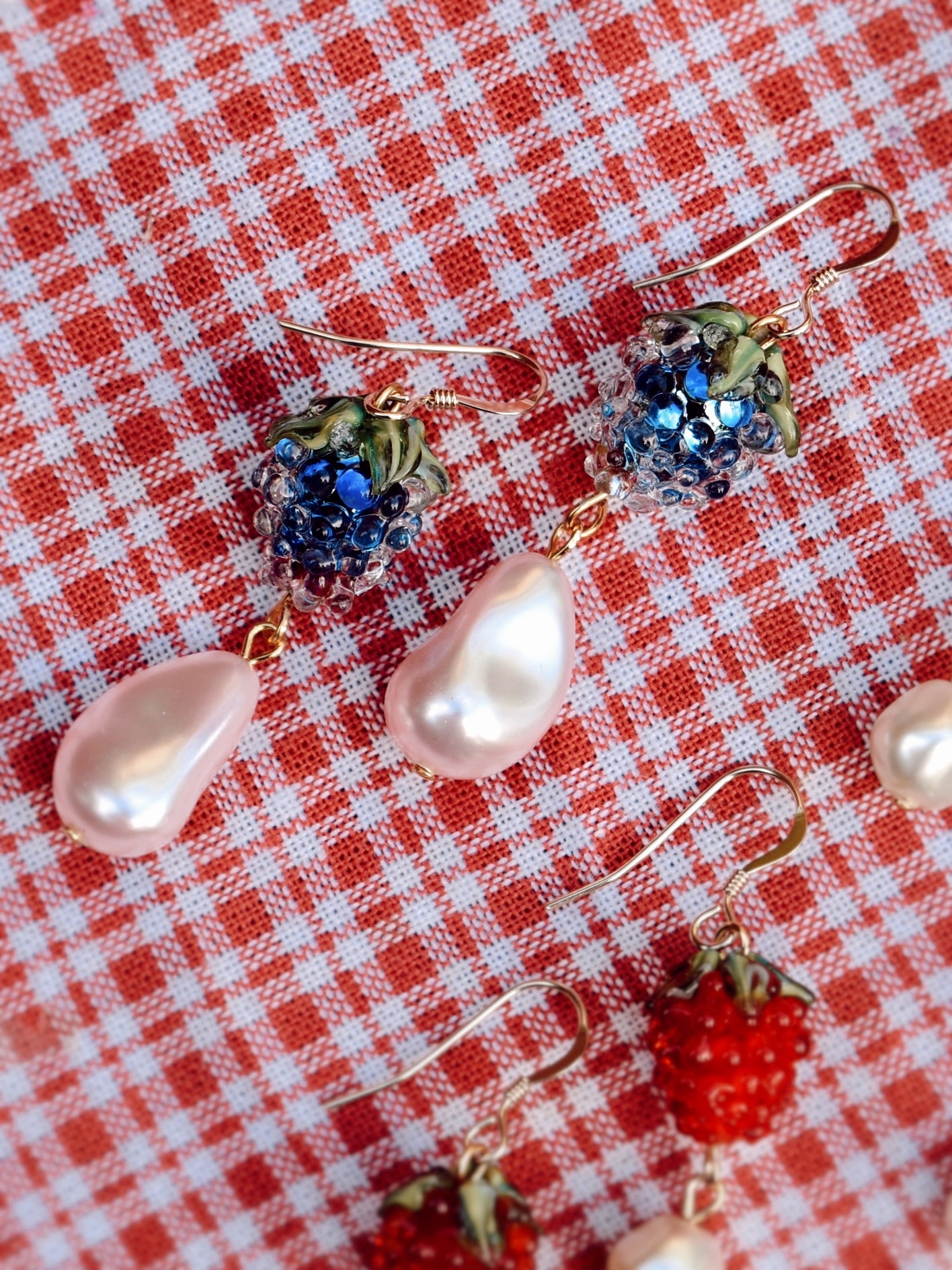 Strawberry-shaped earrings with gemstones and pearls on a red and white checkered background