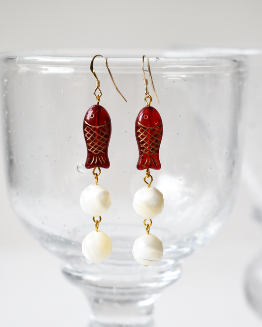 Pair of earrings with red fish-shaped beads and white spherical beads on a glass background