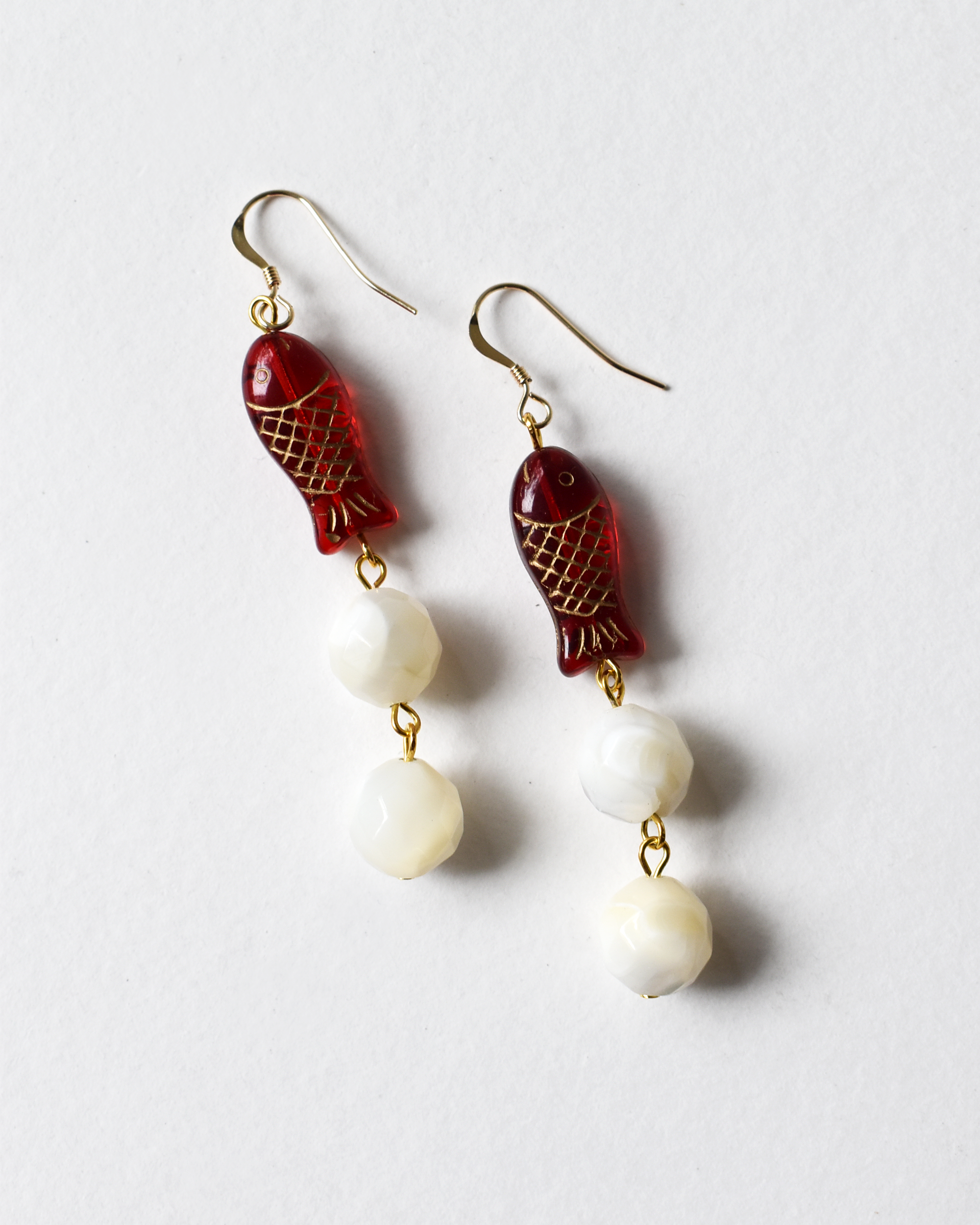 Pair of earrings with red fish-shaped charms and white beads on a light gray background